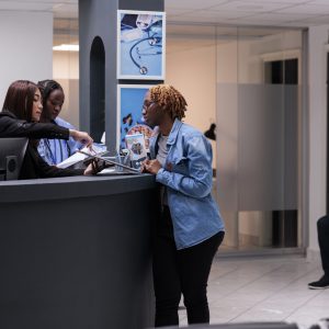 African american woman signing checkup papers at hospital reception desk with receptionist and nurse. Diverse medical team filling in report form files before having consultation appointment.